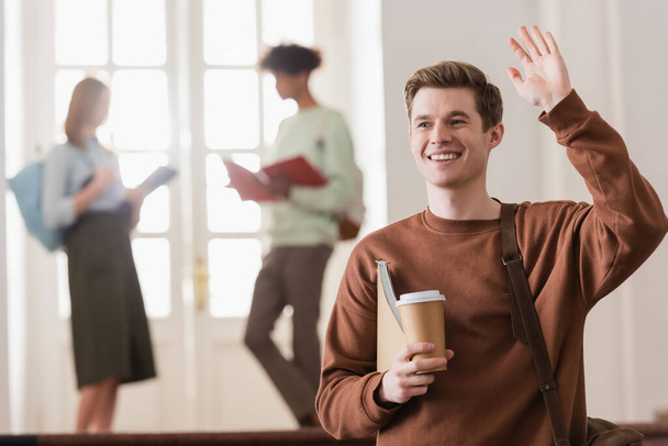 Pozitivní student s kávou jít a notebook mává rukou na univerzitě  - Fotografie, Obrázek