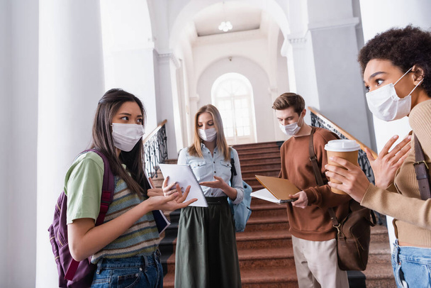 Interracial students with notebooks and paper cup talking in hall of university during quarantine  - Foto, Imagen