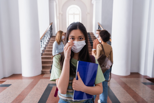 Ασιάτης φοιτητής με ιατρική μάσκα κρατώντας σημειωματάριο στο πανεπιστήμιο  - Φωτογραφία, εικόνα