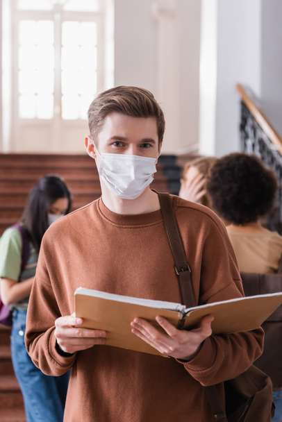 Étudiant en masque médical tenant un carnet sur le premier plan flou à l'université  - Photo, image