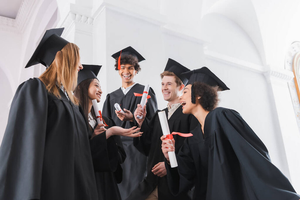 Cheerful interracial graduates in caps holding diplomas  - 写真・画像