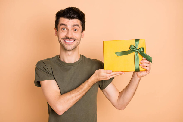 Photo of guy demonstrate present box toothy smile look camera wear green t-shirt isolated beige color background - Zdjęcie, obraz