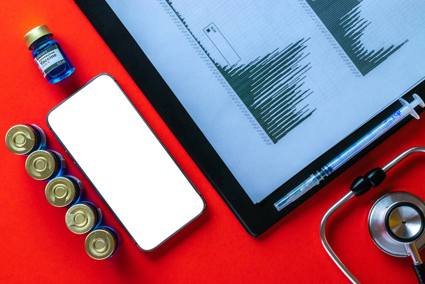 Tıbbi başvuru. Hastane ekipmanları: doktor stetoskobu, sağlık çizelgeleri, iğneli şırınga ve kırmızı arka planda boş ekranlı siyah akıllı telefon. - Fotoğraf, Görsel