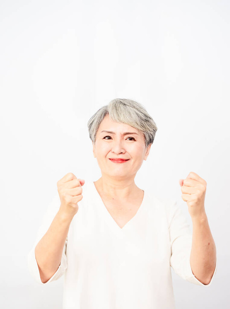Senior beautiful asian woman wearing casual dress standing and raising fists in white background. - Photo, Image