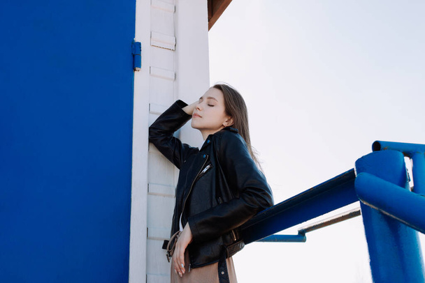 Jonge vrouw in zwart leren jasje buigt over leuningen voor een witte muur, blauwe deur. Veel plezier buiten. Stedelijke stijl en straatmode. Meisje met gesloten ogen in casual outfit aanraken van haar - Foto, afbeelding