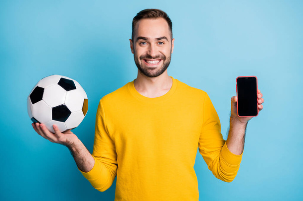 Photo of young handsome happy positive cheerful man show phone touchscreen hold football ball isolate on blue color background - Zdjęcie, obraz
