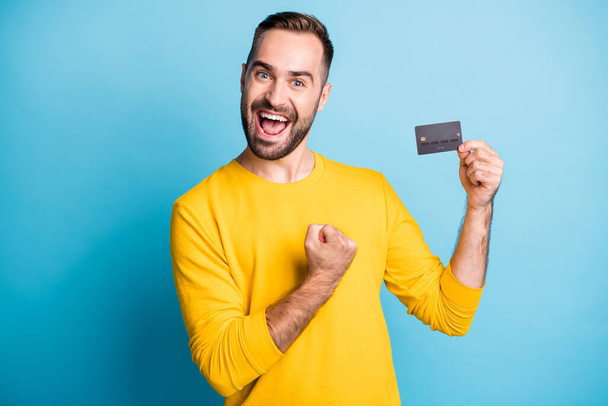 Photo of young excited happy positive cheerful smiling man hold credit card raise fist in victory isolated on blue color background - Fotó, kép