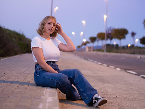 A blonde Caucasian woman from Spain in a white crop top sitting on a sidewalk curb - Foto, Imagen