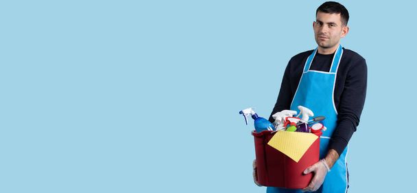 Banner, long format on blue background. Young serious caucasian man carrying a bucket with diverse cleaning products. Man doing home work, home life, cleans dirty room with washing products - Photo, image