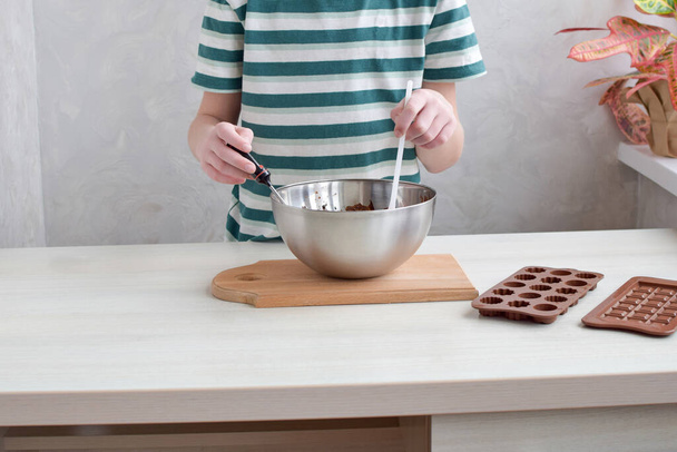 Schritt für Schritt bereitet der Junge zu Hause handgemachte Schokolade zu. Flüssige Schokolade mit einem Spachtel auf einem hellen Tisch verrühren. - Foto, Bild