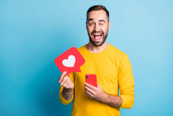 Photo of young happy excited smiling cheerful man wink eye hold phone and heart icon isolated on blue color background - Foto, afbeelding