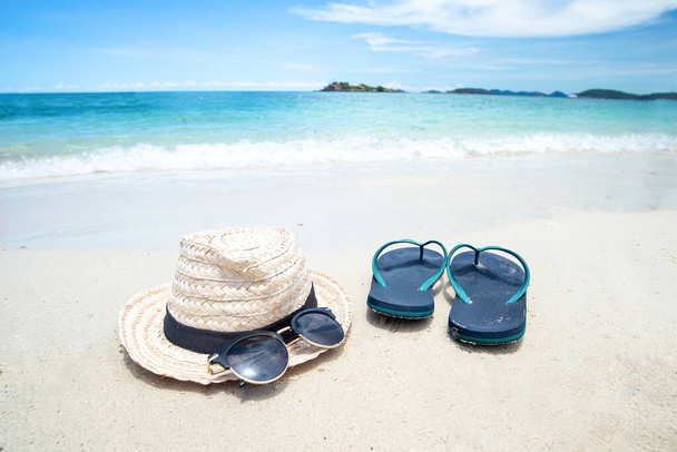 新鮮な海の背景にスリッパの近くに麦藁帽子と黒サングラス。夏、旅行や休暇のコンセプトとして美しい砂浜。休日の概念。メッセージのコピースペース. - 写真・画像