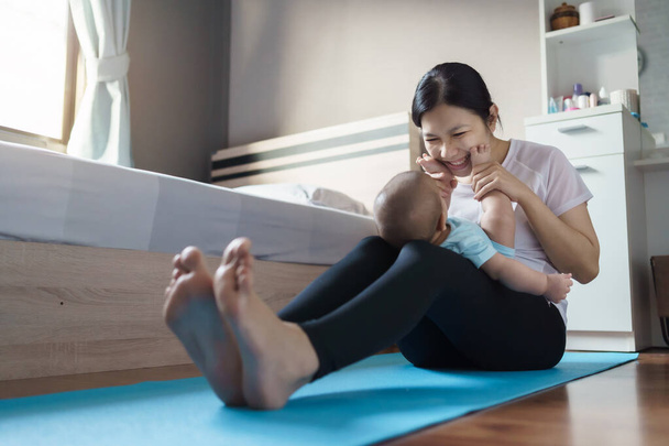 Happy smiling Asian young mother in sportswear playing and holding small baby son feet while practicing yoga in bedroom at home. - Fotó, kép