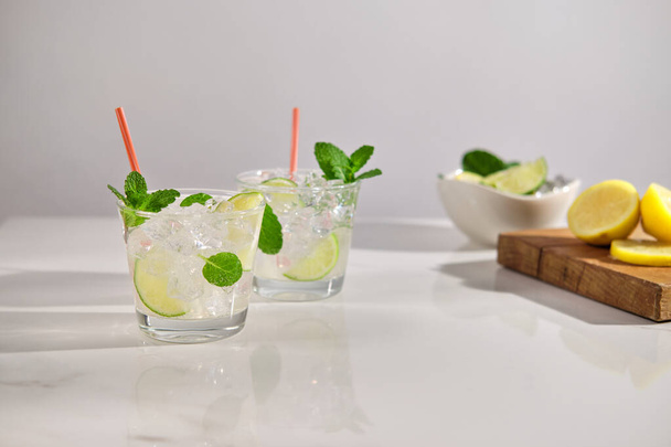 Verres de cocktail Mojito avec glaçons, menthe et citron vert sur marbre blanc avec citrons verts frais, citrons et menthe. Cocktail rafraîchissant pétillant. Espace pour le texte - Photo, image