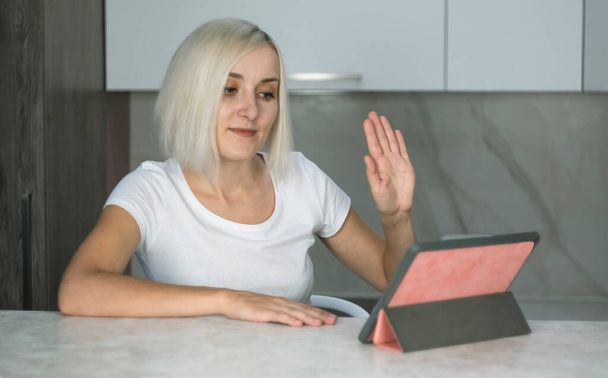 Schöne junge blonde Frau spricht per Videoanruf, während sie auf den Bildschirm des Tablets schaut. Erhob ihre Hand, grüßt den Gesprächspartner - Foto, Bild