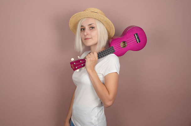Hermosa joven rubia en sombrero de paja con ukelele sobre fondo rosa - Foto, Imagen