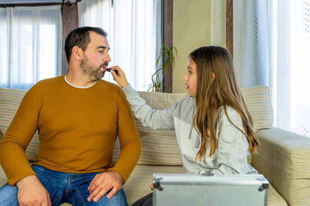 Beautiful pre-adolescent girl painting her father's lips in the living room at home. Fun concept - Zdjęcie, obraz