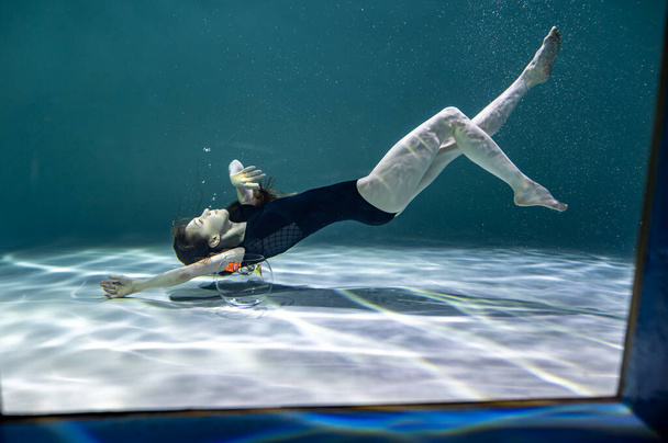 красивая девушка в черном купальнике играет с рыбой в аквариуме на синем фоне под водой - Фото, изображение