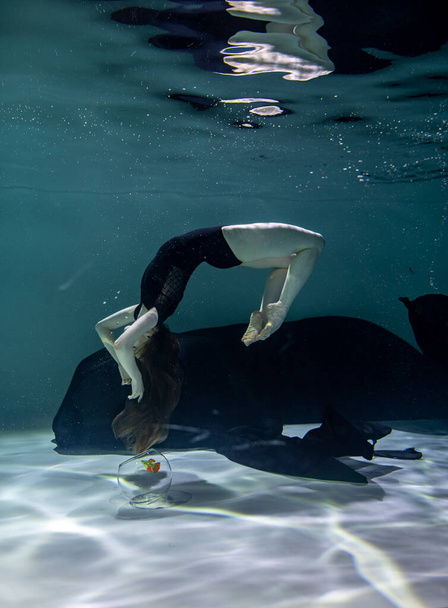 mooi meisje in een zwart badpak met een zwarte pareo speelt met vissen in een aquarium op een blauwe achtergrond onderwater - Foto, afbeelding