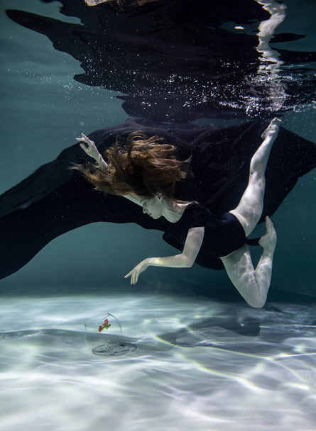 όμορφο κορίτσι με μαύρο μαγιό με μαύρο παρεό παίζει με ψάρια σε ενυδρείο σε μπλε φόντο κάτω από το νερό - Φωτογραφία, εικόνα