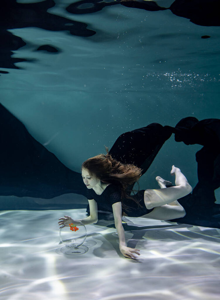 όμορφο κορίτσι με μαύρο μαγιό με μαύρο παρεό παίζει με ψάρια σε ενυδρείο σε μπλε φόντο κάτω από το νερό - Φωτογραφία, εικόνα