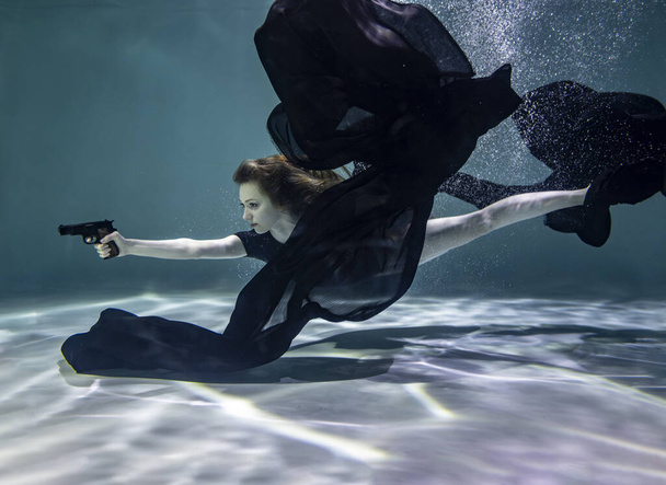 piękna dziewczyna w czarnym stroju kąpielowym z czarnym pareo trzyma czarny pistolet w rękach na niebieskim tle pod wodą - Zdjęcie, obraz