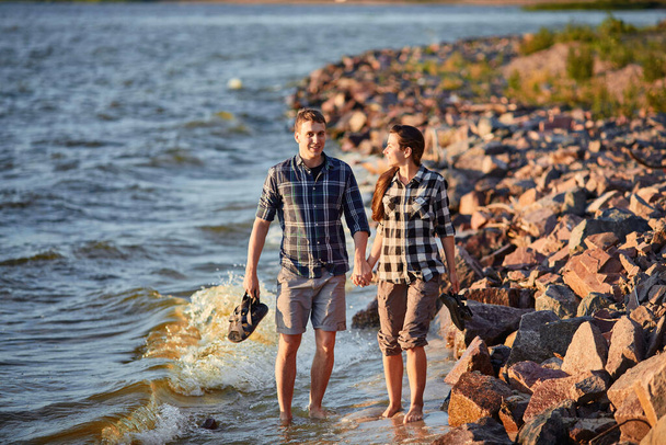 Ευτυχισμένο καυκάσιο μοντέρνο ζευγάρι στην αγάπη κρατώντας τα χέρια και περπατώντας στην ακτή κοντά στο ποτάμι. Στο βάθος είναι το ηλιοβασίλεμα. - Φωτογραφία, εικόνα