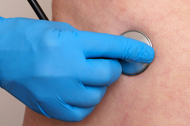 La main d'un médecin dans un gant bleu tient un stéthoscope sur le corps du patient. Gros plan. - Photo, image