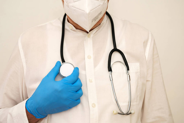 A stethoscope on the chest of a masked doctor and a hand in a blue latex glove. Closeup. - Fotografie, Obrázek