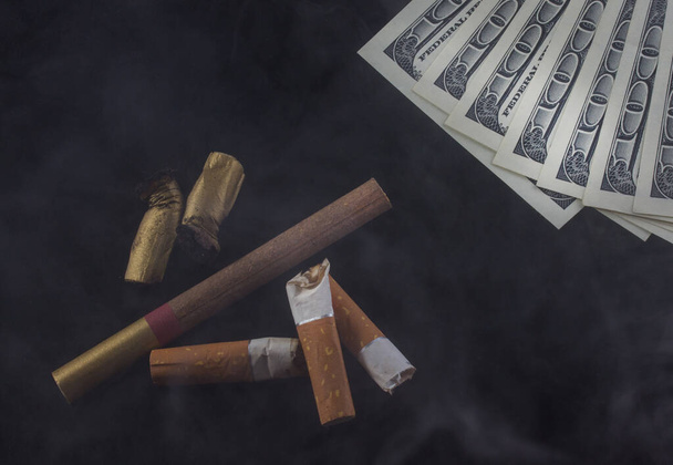 mozziconi di sigaretta e banconote americane da cento dollari si trovano nel fumo su uno sfondo scuro il concetto del costo del fumo e della droga - Foto, immagini