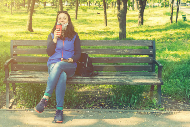 Beautiful Caucasian woman with long brown hair sitting in the park drinking a cup of tea or coffee and smiling. Middle-aged athletic woman with long hair drinks coffee in a park. toned - Photo, image