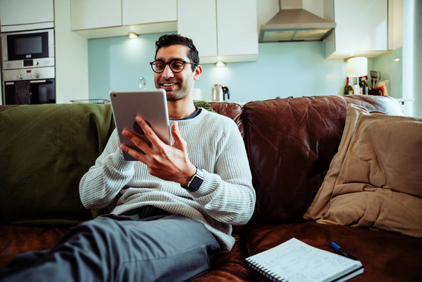 Glücklicher gemischter Geschäftsmann, der von zu Hause aus auf dem digitalen Tablet tippt und auf der gemütlichen Couch in der Lounge sitzt - Foto, Bild