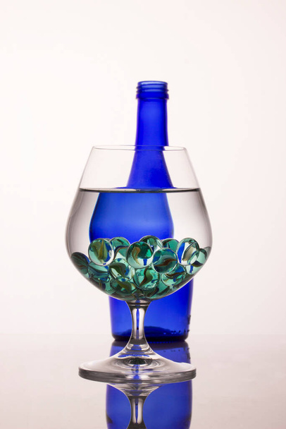 Dekorative Glaskugeln liegen auf dem Boden eines Glases mit Wasser gegen eine blaue Flasche, die verzerrt ist - Foto, Bild