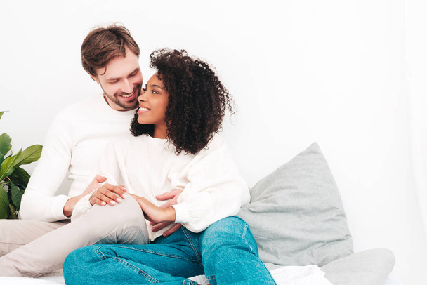 Smiling beautiful woman and her handsome boyfriend. Happy cheerful multiracial family having tender moments. Multiethnic models lying in bed and hugging in white interior. Embracing each other - 写真・画像