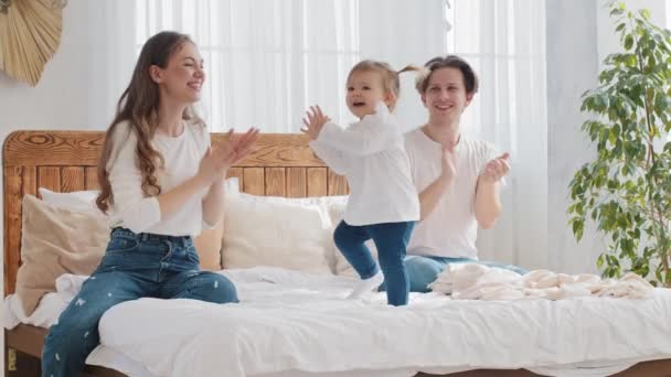Boldog család fiatal szülők a baba kislány csecsemő ül az ágyon, kislány kisgyerek táncol a zene anya és apa tapsol tapsol kéz támogatja a gyermek tánc, szórakoztató szabadidő otthon hálószoba - Felvétel, videó