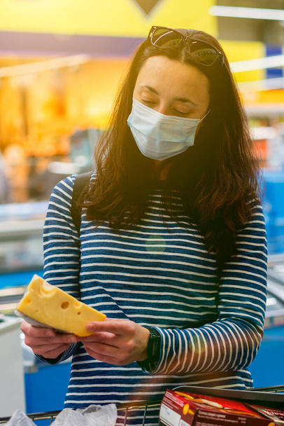 woman in protective medical mask in a supermarket chooses cheese. pandemic and covid-19 concept. Hand of the buyer with a piece of cheese in the store. vertical photo - Photo, Image