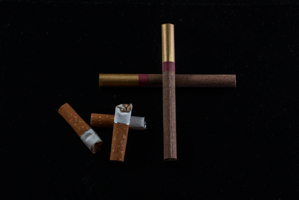 krzyż wyłożony z papierosów i niedopałków papierosów leży obok na ciemnym tle pojęcia palenia i złych nawyków - Zdjęcie, obraz