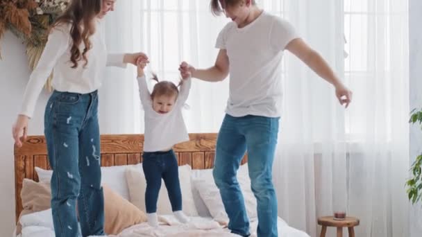 Mladí milující běloši rodiče hrají doma baví si herní čas s malou dceruškou dítě v posteli v ložnici, matky a otcové drží dítě malé holčičky rukou zvednout dítě ve vzduchu - Záběry, video