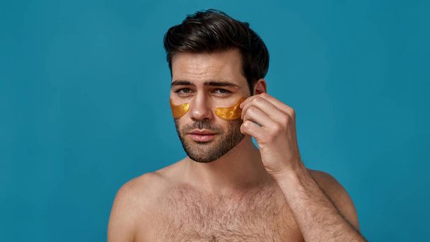Good looking brunette guy applying golden gel under eye patches on his skin while looking at camera, posing isolated over blue background - Foto, imagen