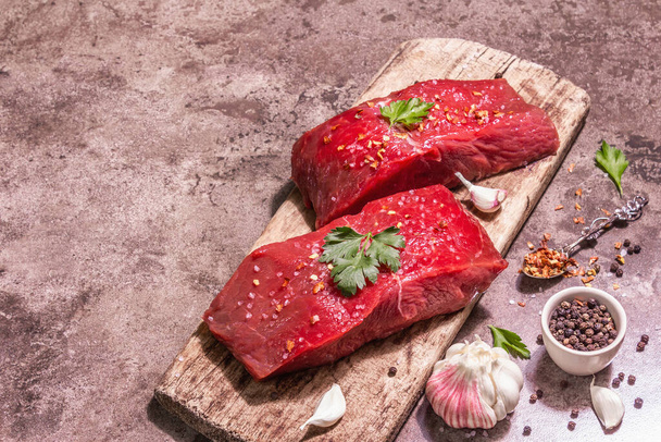 Raw beef steaks on a wooden cutting board. Trendy hard light, dark shadow. Spices, fresh garlic, and parsley. Stone concrete background, top view - Photo, Image