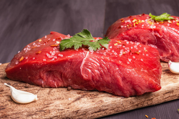 Raw beef steaks on a wooden cutting board. Spices, fresh garlic, and parsley. Black wooden boards background, close up - Φωτογραφία, εικόνα