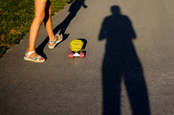 公園のパス上のペニースケートボードを持つ子供に焦点を当てます。男の子と女の子は太陽の日にペニーボードに乗る。晴れた夏の日に屋外のライフスタイル写真。長い影だ。趣味がいい。集中できない. - 写真・画像