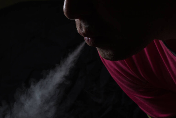 Der dünne, graue Dampf einer elektronischen Zigarette kommt aus dem Mund einer rauchenden Person auf dunklem Hintergrund, der an schlechte Angewohnheit und elektronische Zigaretten erinnert. - Foto, Bild