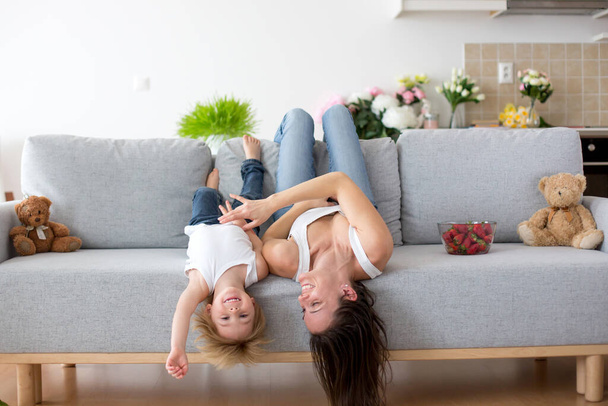 Mother and toddler child, hanging upside down from a couch at home, smiling happily - Foto, Imagem