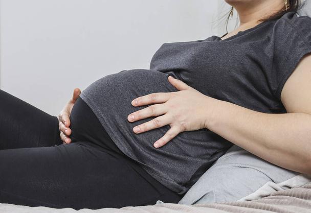 Una mujer embarazada sostiene su vientre con las manos, acostada en la cama, vista lateral de cerca. - Foto, imagen