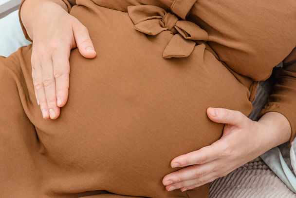 Eine schwangere Frau hält ihren Bauch mit den Händen auf dem Bett liegend, von oben gesehen, Nahaufnahme. - Foto, Bild