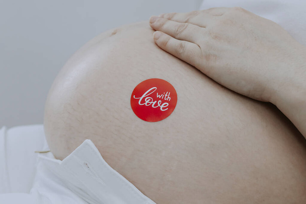 Una mujer embarazada sostiene su vientre con su mano con una pegatina roja: con amor, vista lateral de cerca. - Foto, imagen