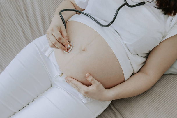 Беременная женщина, лежащая на кровати, слушает стетоскопом свой живот, вид сверху крупным планом. - Фото, изображение