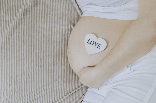 Μια έγκυος γυναίκα κρατά την κοιλιά της με μια ξύλινη καρδιά, που βρίσκεται στα δεξιά στο κρεβάτι και με μια θέση για κείμενο στα αριστερά, πάνω άποψη γκρο πλαν. - Φωτογραφία, εικόνα