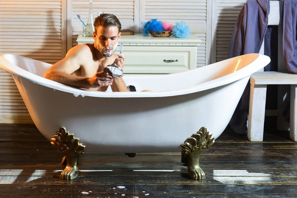 Shaving man. Guy shave with razor in bath in bathroom. Macho with shaving soap on beard hair look in mirror - Foto, immagini